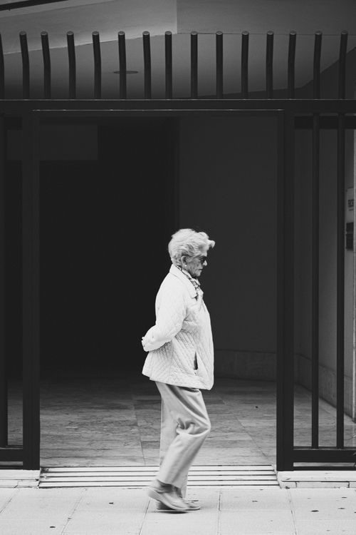 Free Black and white photo of a senior woman walking on a city sidewalk, showcasing fashion. Stock Photo
