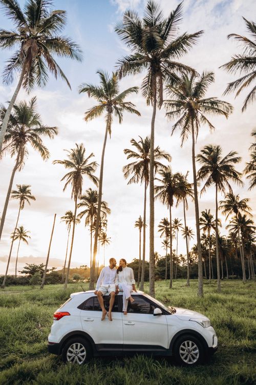 Free A couple relaxes on a car roof amidst tall palm trees during a serene tropical sunset. Stock Photo