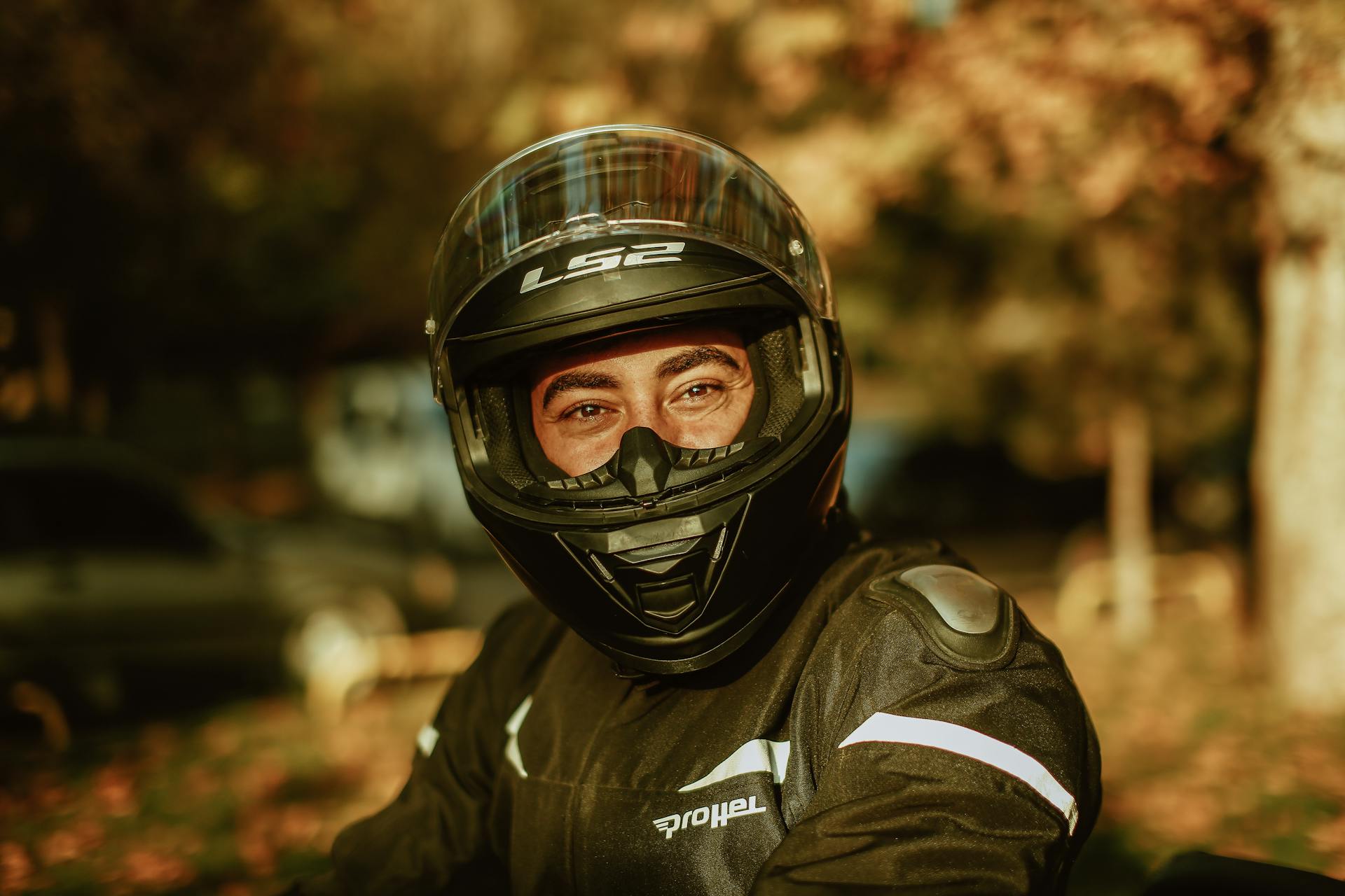 Close-up of a motorcyclist wearing protective gear and helmet outdoors in autumn light.