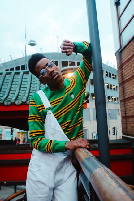 Stylish man in green striped sweater and overalls poses in urban landscape.