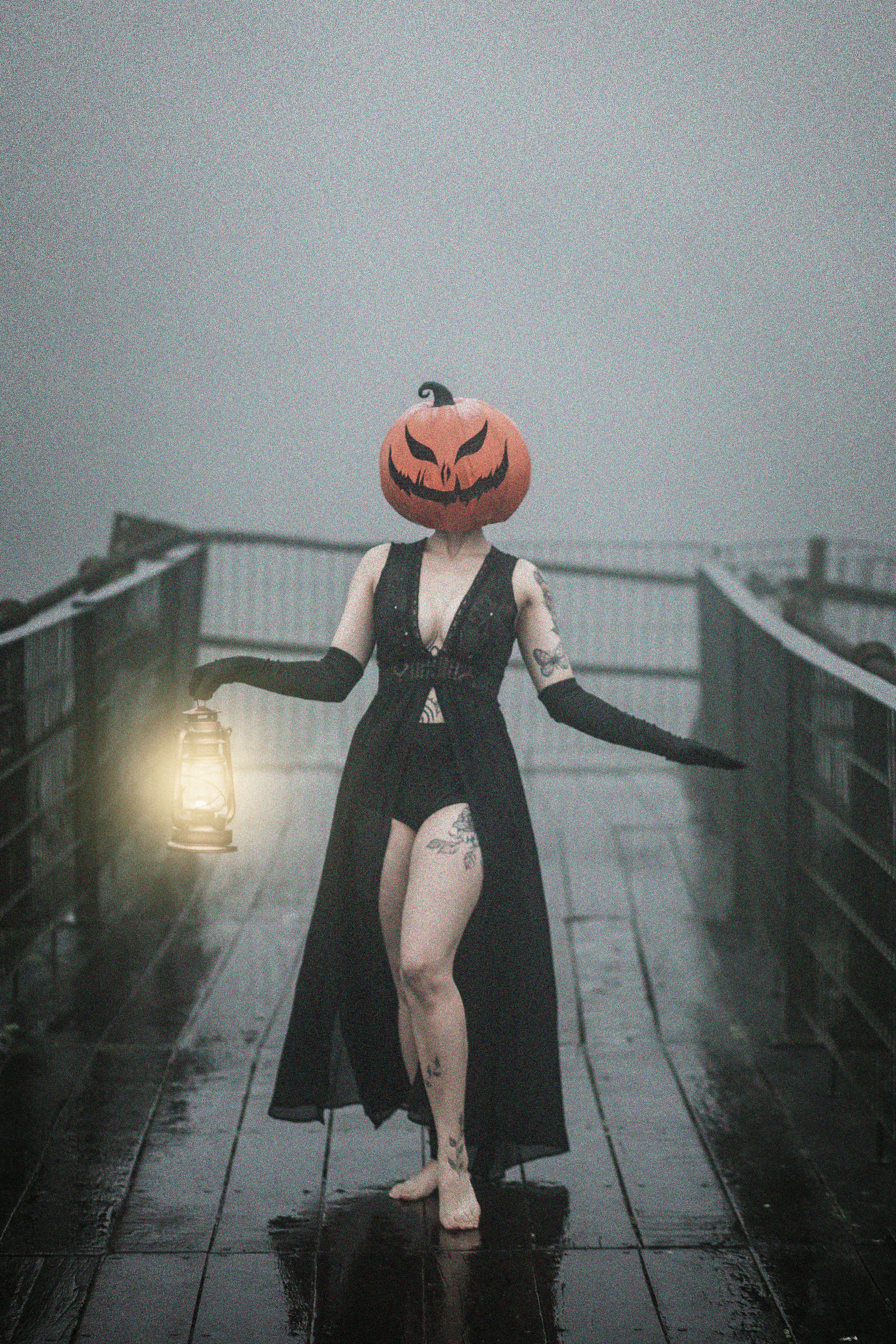 Eerie scene of a woman with a pumpkin head holding a lantern on a foggy bridge. Perfect for Halloween themes.
