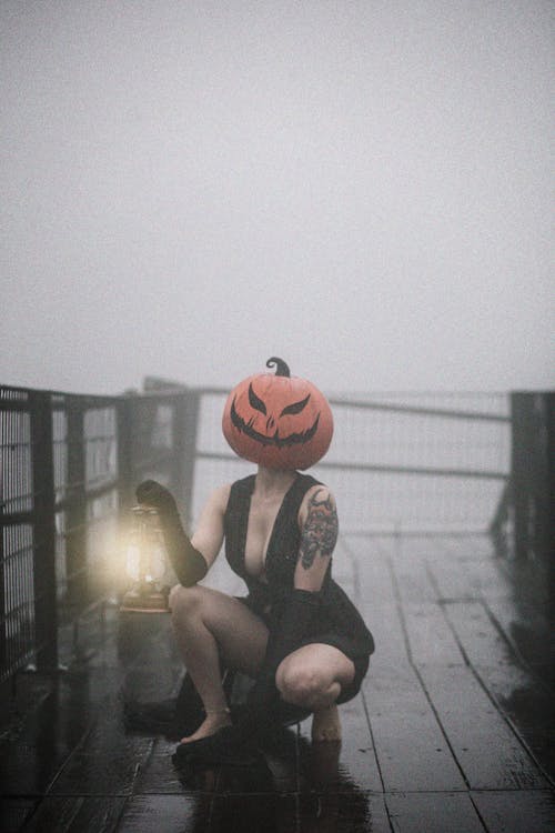 Woman in Black Dress with Pumpkin Mask Crouching on Pier