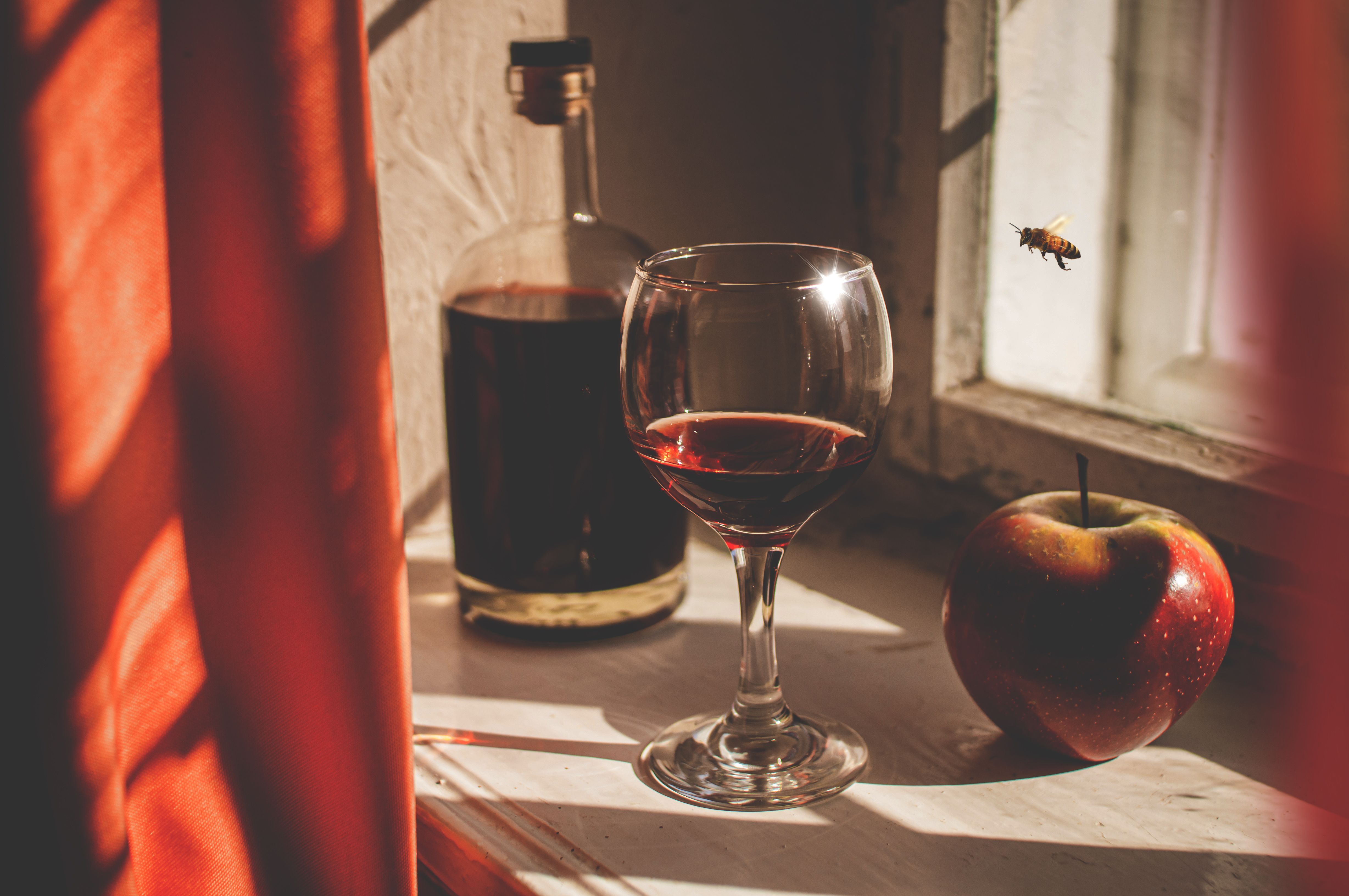 A glass of red wine, apple, and bottle on a sunlit windowsill with a bee hovering.