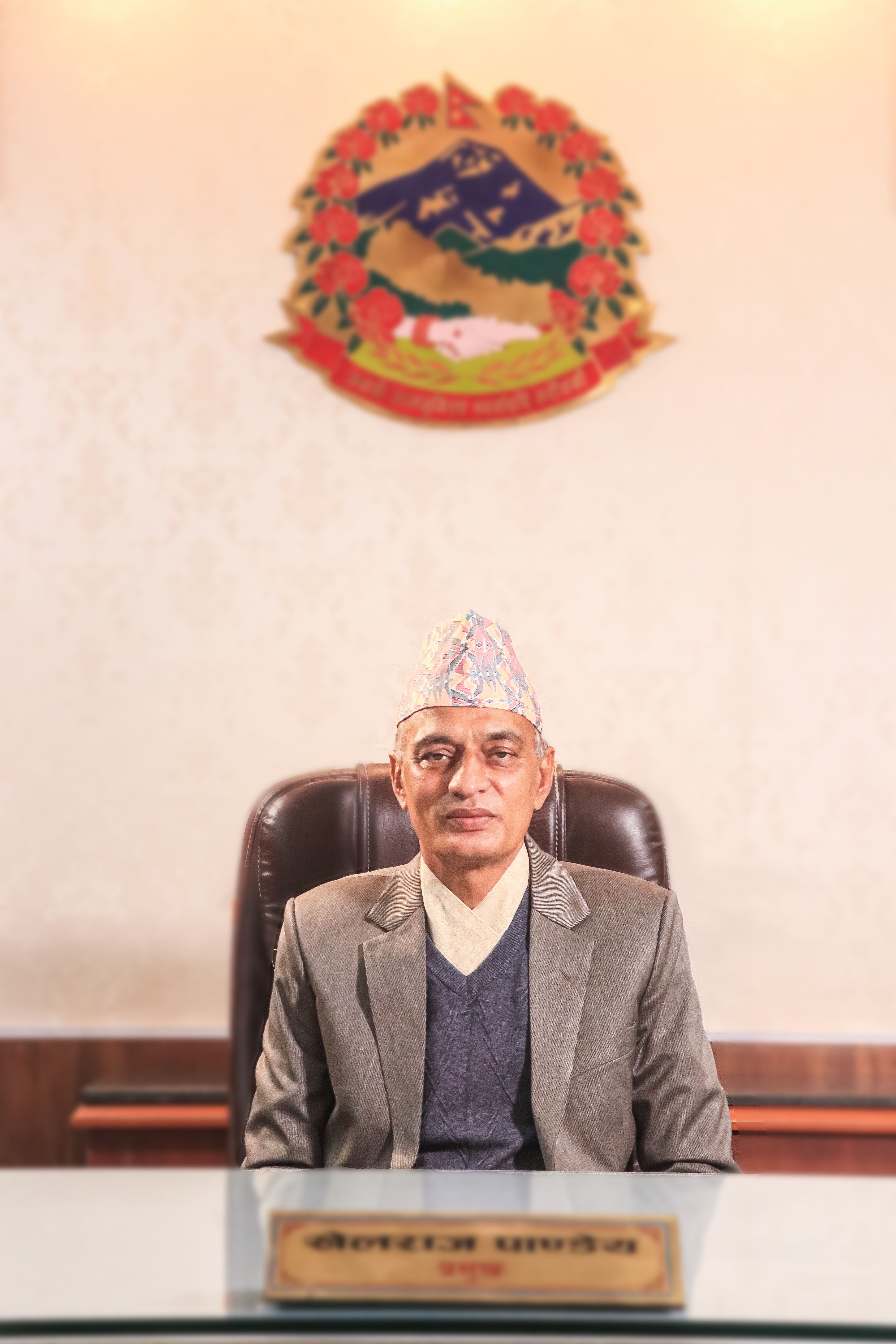 Portrait of a senior official in a Nepali government office, seated with an emblem on the wall.
