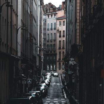 A moody city street framed by historical buildings and parked cars in a narrow urban setting.