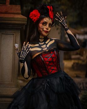 Woman in Day of the Dead makeup and traditional attire poses in a cemetery setting, showcasing cultural heritage.