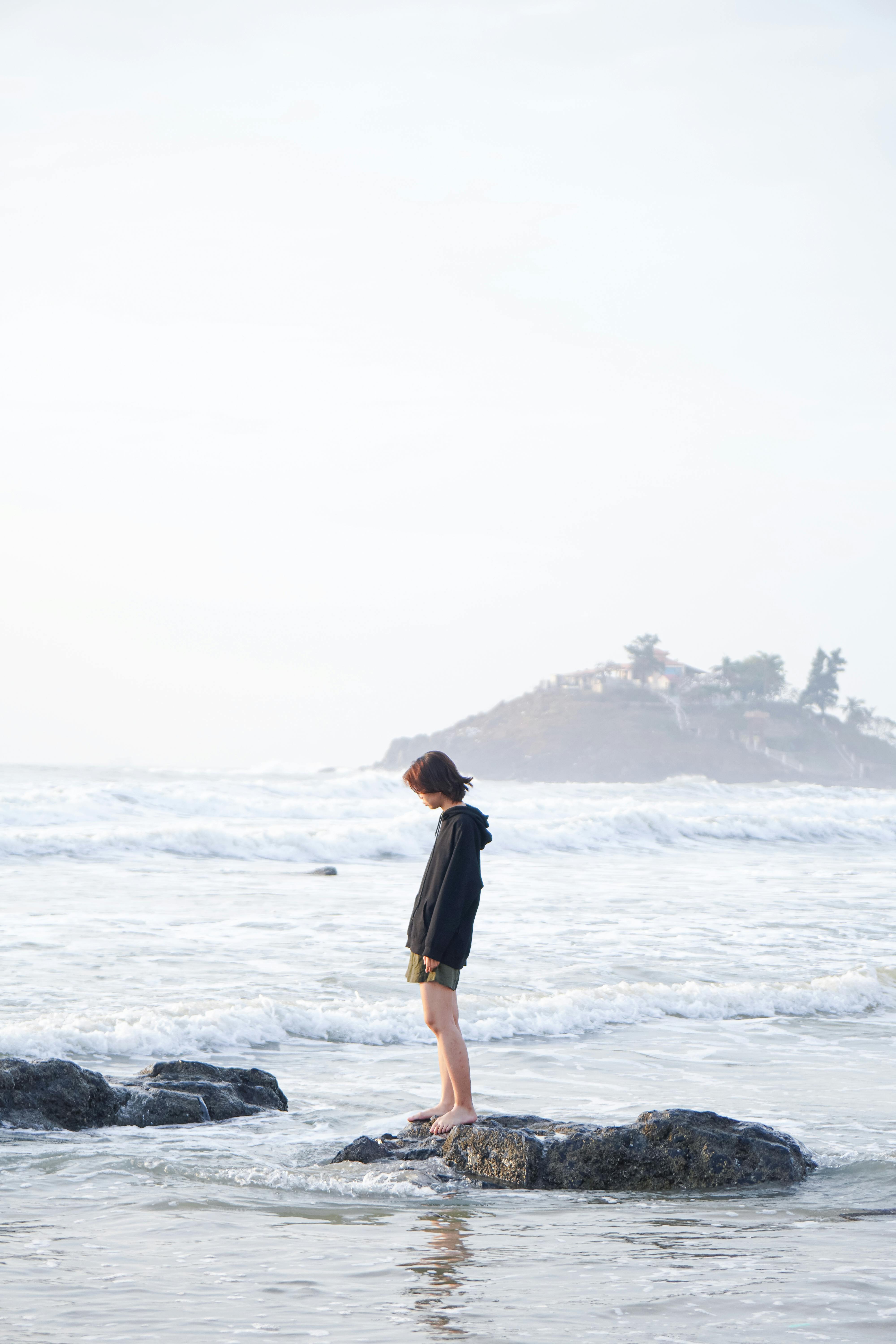 A person stands thoughtfully on a rock by the sea with gentle waves in the background.