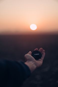 A person holds a camera lens cap with a beautiful sunset in the background