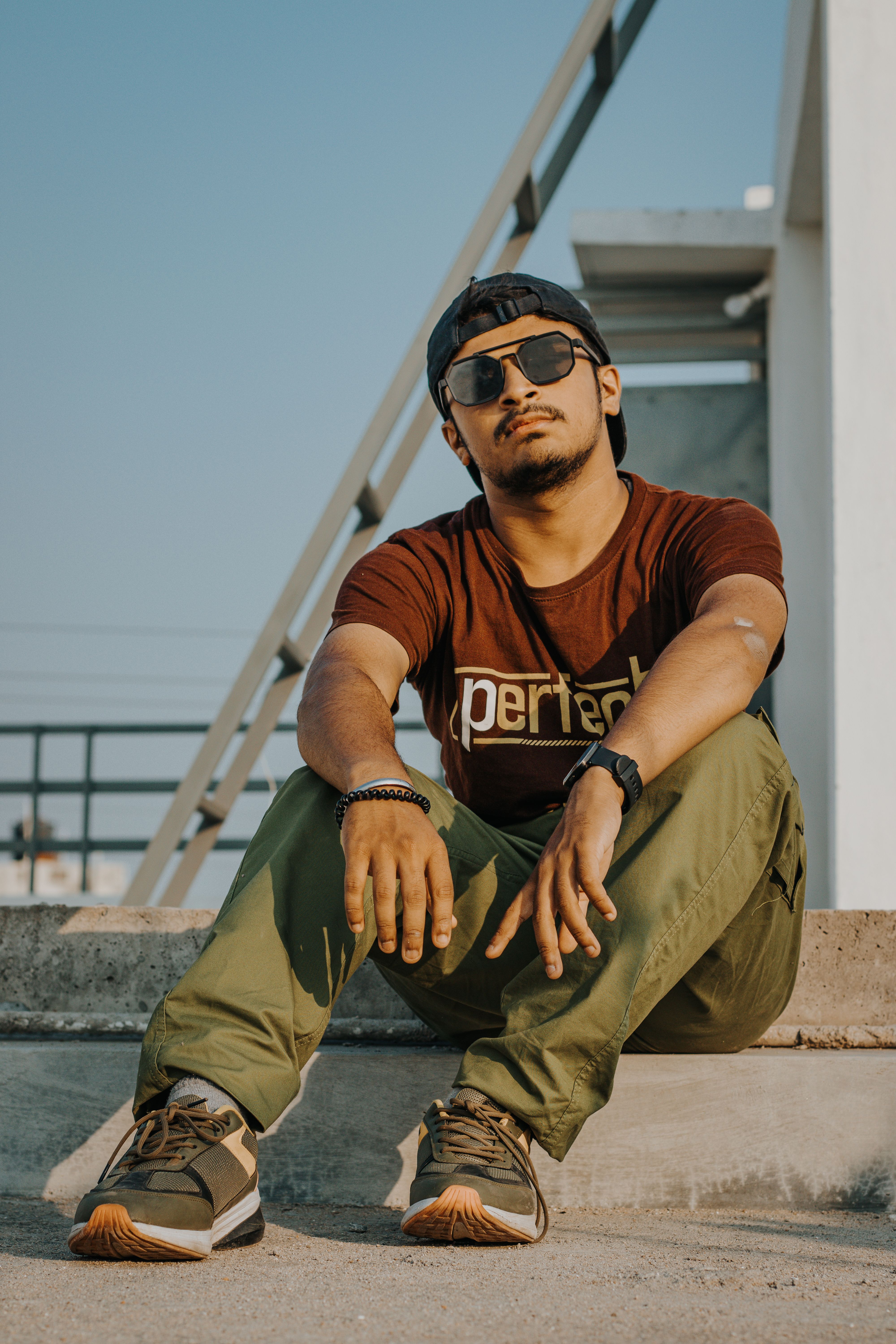 Stylish young man sitting on rooftop wearing sunglasses and casual attire, showcasing urban fashion.