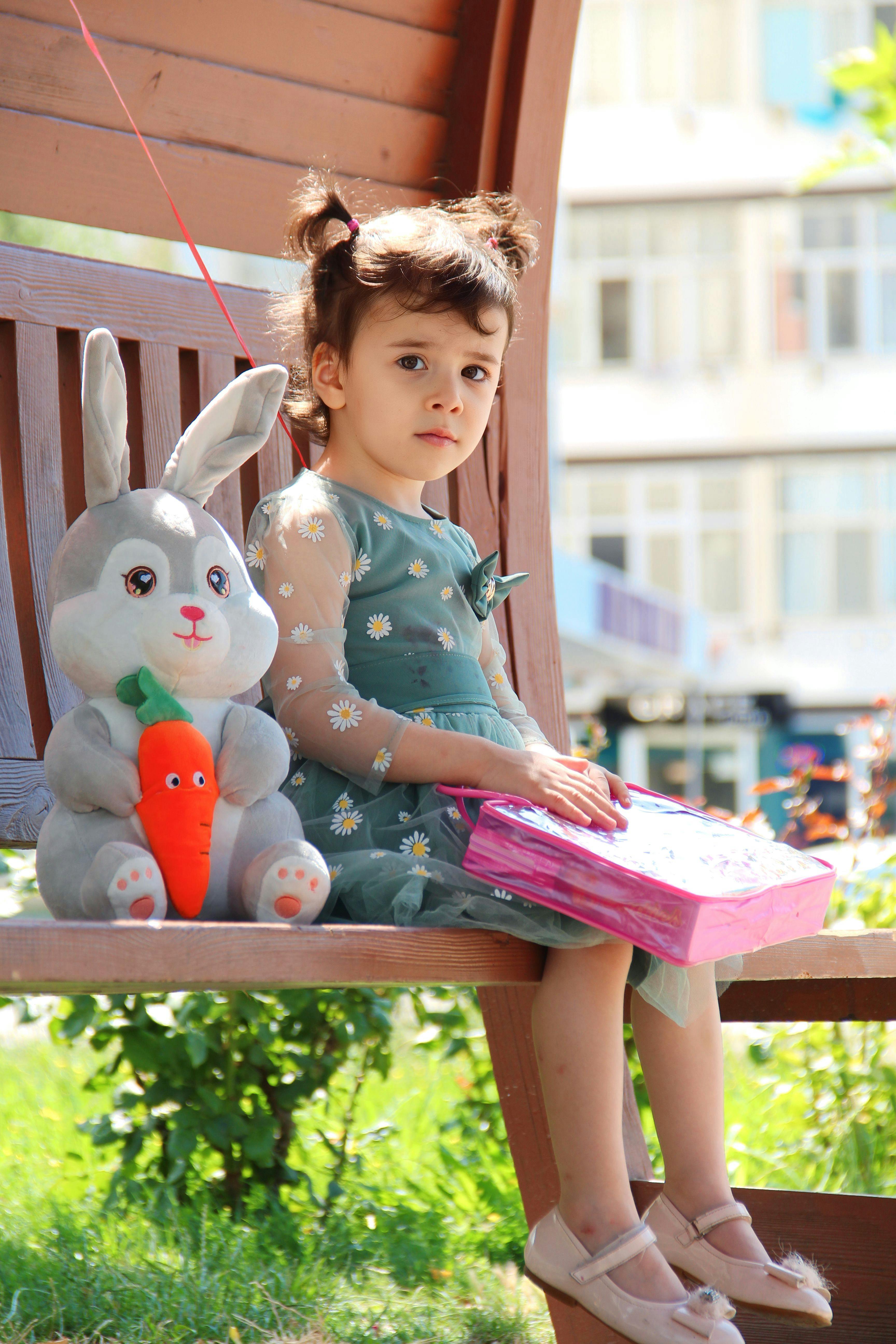 Little girl sitting with toy rabbit and book on a sunny park bench.