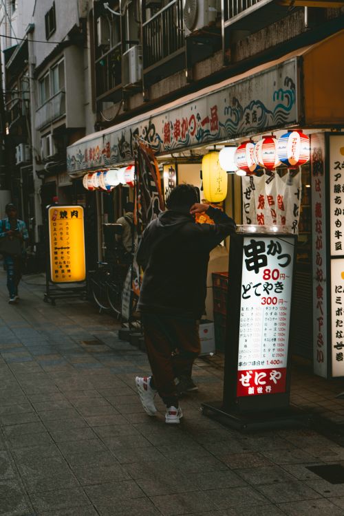 Free A person leans on a Japanese restaurant board in a bustling urban street at night. Stock Photo