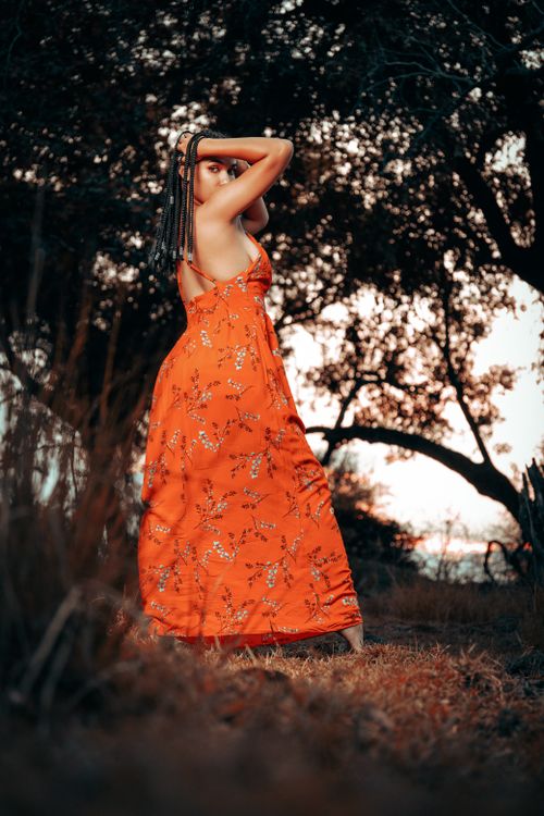 Free A woman in a floral orange dress stands outdoors, surrounded by trees during sunset. Stock Photo