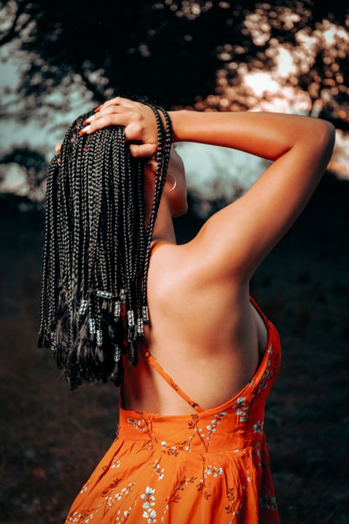 Free Back view of a woman with braided hair enjoying a sunset outdoors in a bright orange summer dress. Stock Photo