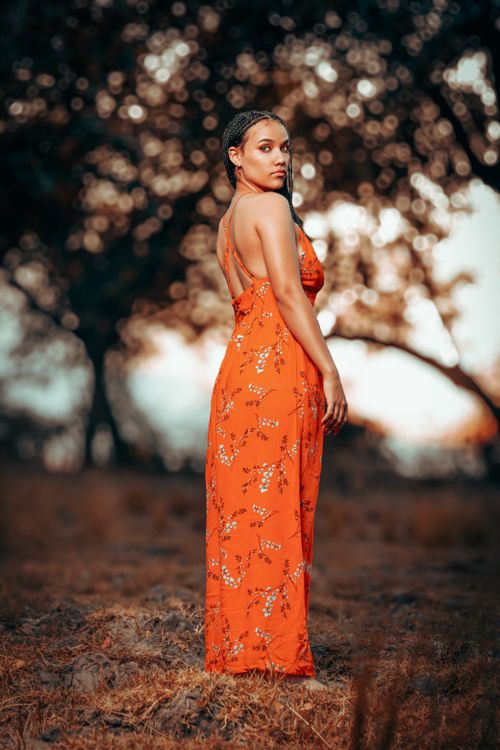Free A young woman in a floral orange dress poses gracefully outdoors at sunset. Stock Photo