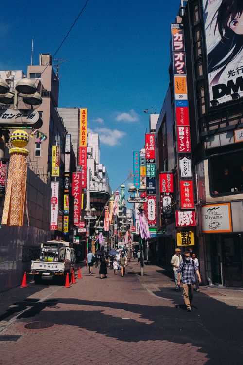 Free Bustling Tokyo street with neon signs and walking pedestrians under a bright blue sky. Stock Photo