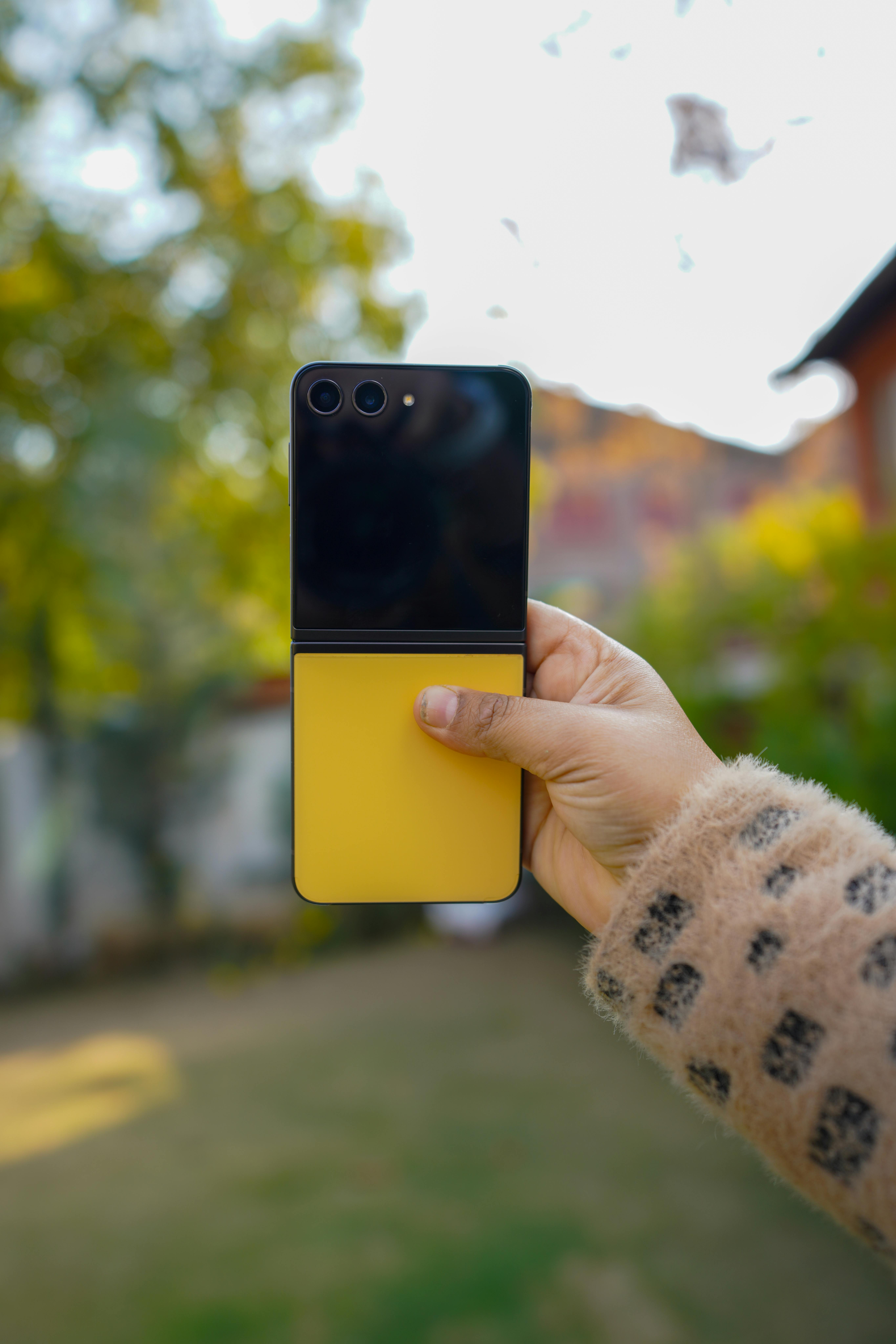 Free Close-up of a hand holding a yellow smartphone outdoors in a sunny backyard. Stock Photo