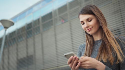 Free Young woman smiling while using a smartphone outside in an urban area. Stock Photo