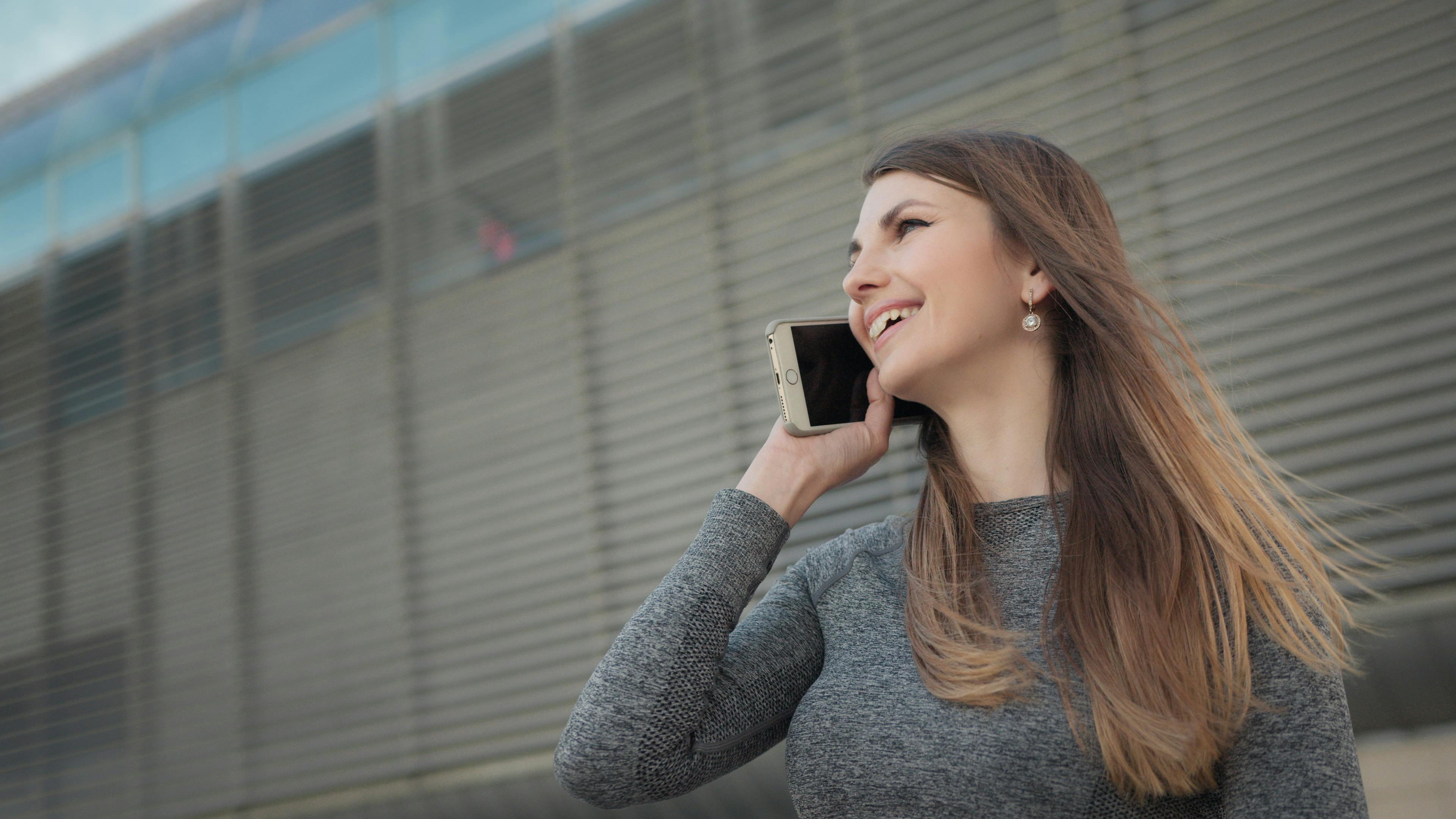 Woman in urban setting talking on phone, wearing casual attire, smiling.