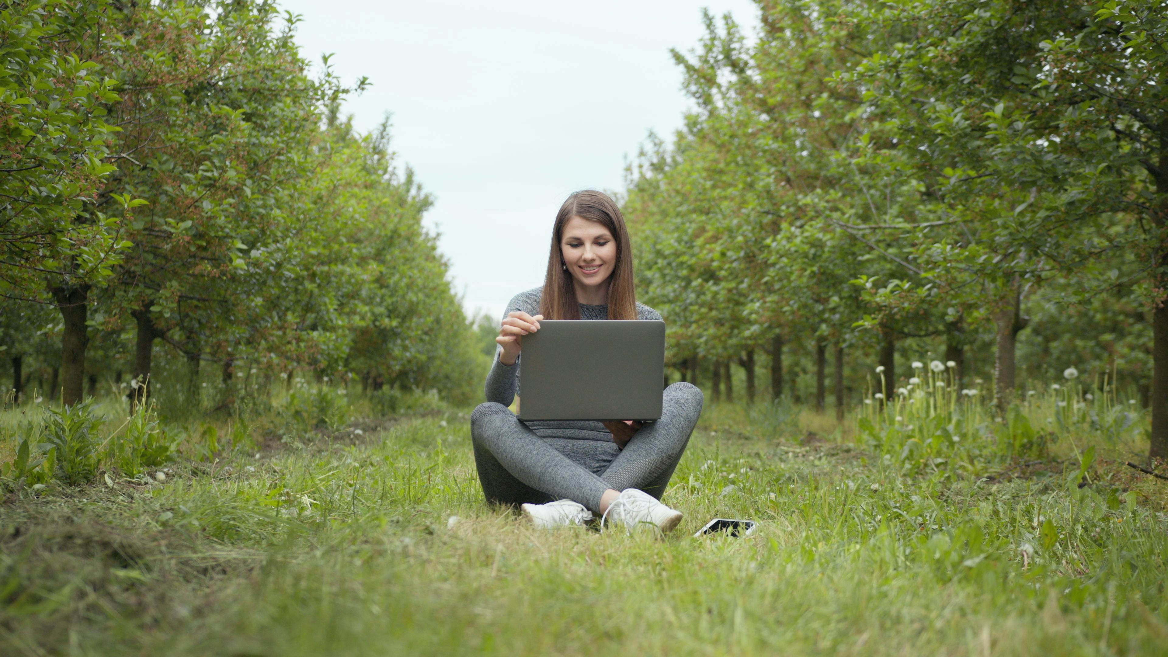 Young woman sits with laptop in an orchard, enjoying a peaceful work environment.