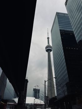 Dramatic view of Toronto's CN Tower amidst towering modern skyscrapers on a cloudy day.