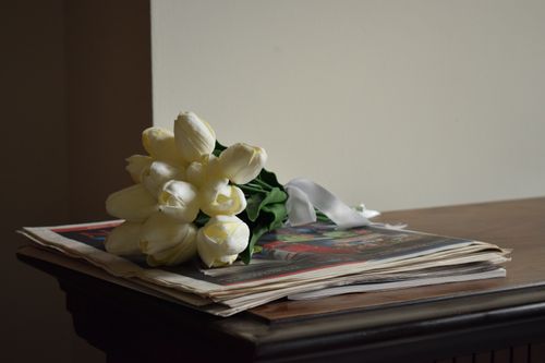 Free A graceful arrangement of white tulips rests on a stack of newspapers on a vintage wooden table. Stock Photo