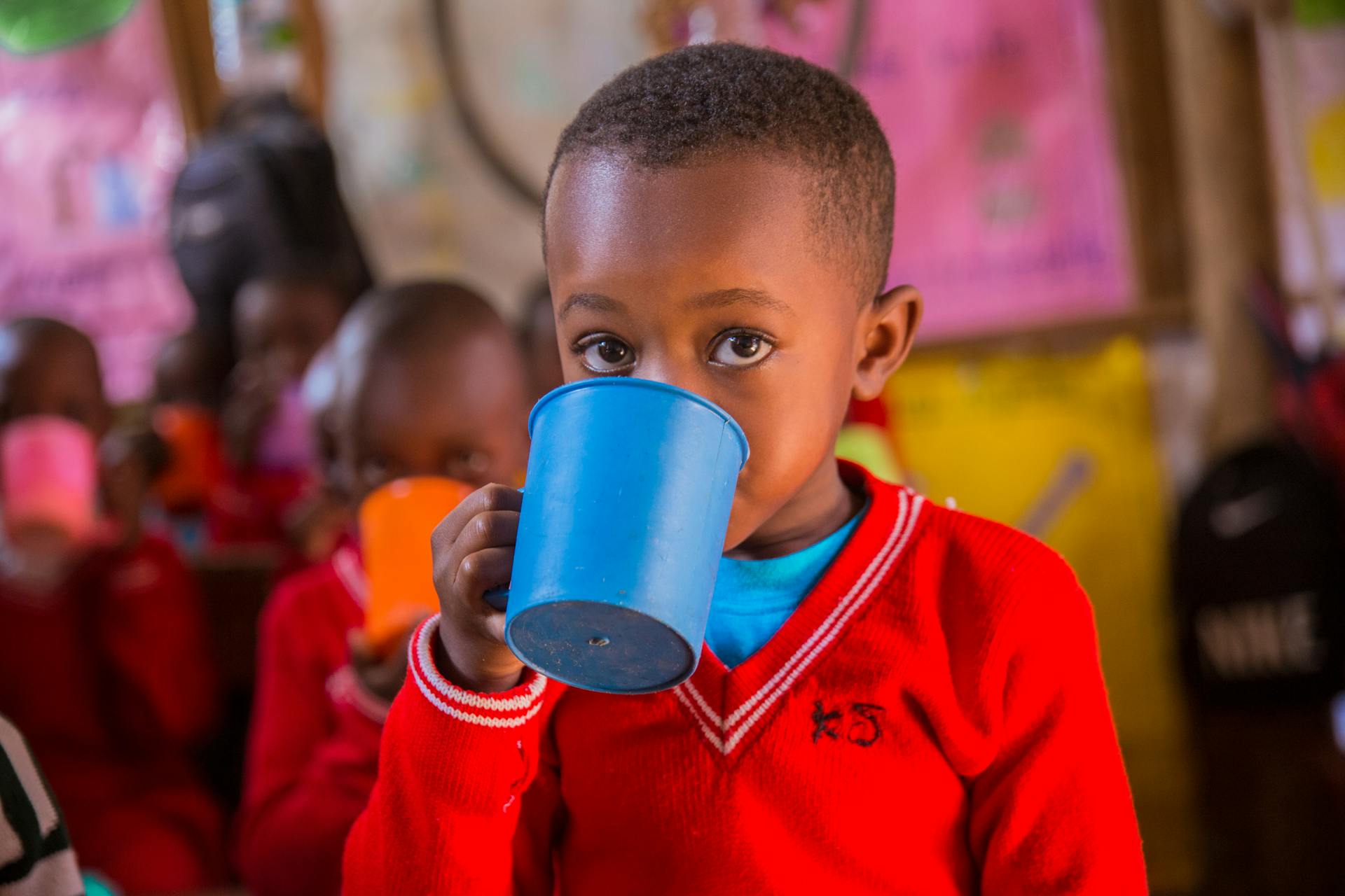 A young child in a vibrant Ugandan classroom enjoys a drink from a colorful cup.