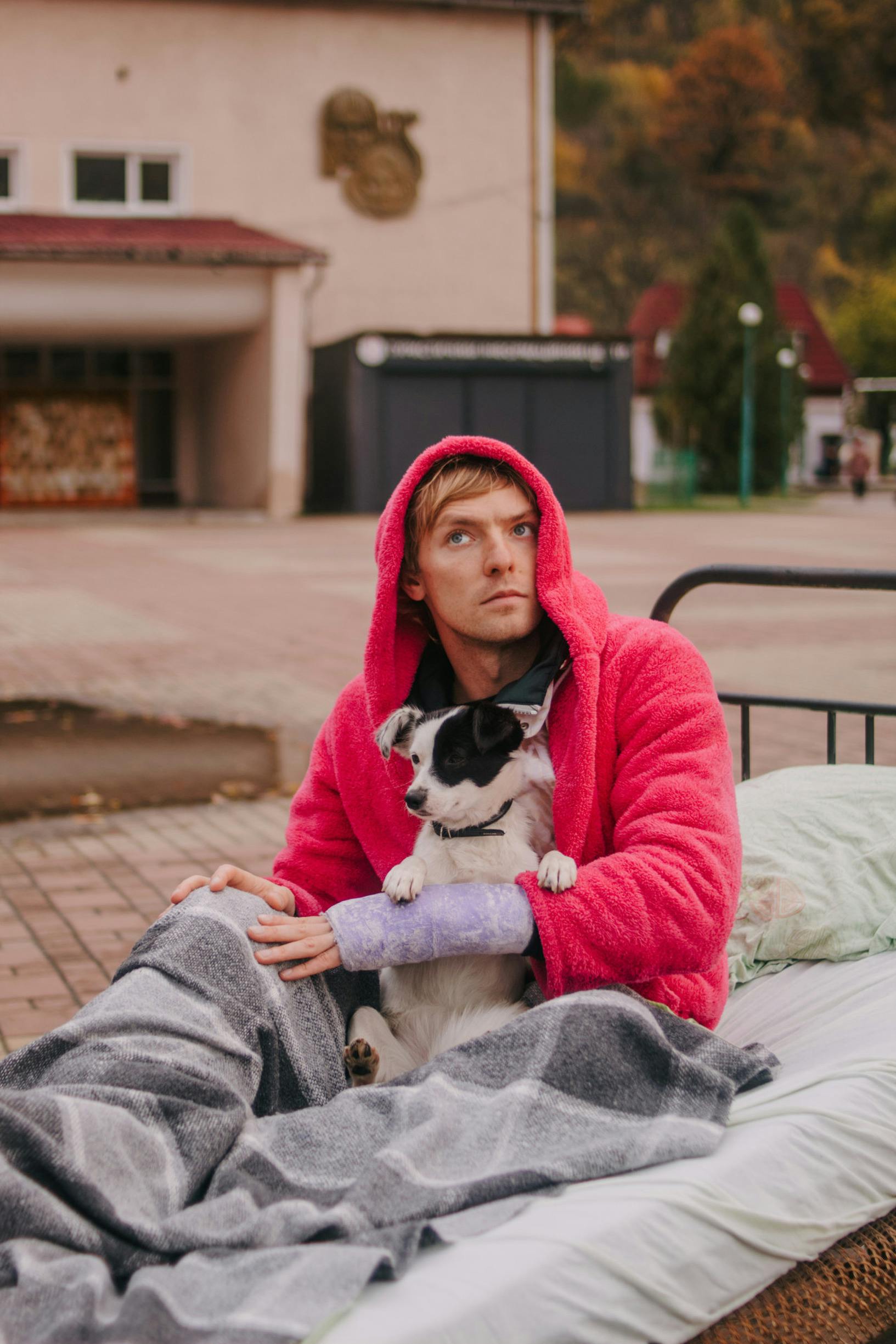 Man in pink hoodie sits outdoors with a dog on a bed in a cozy, autumn setting.