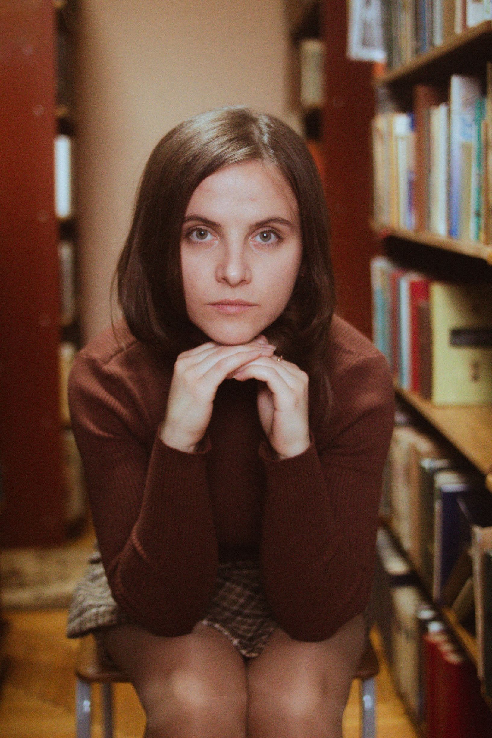A contemplative young woman sits in a cozy library setting with bookshelves.