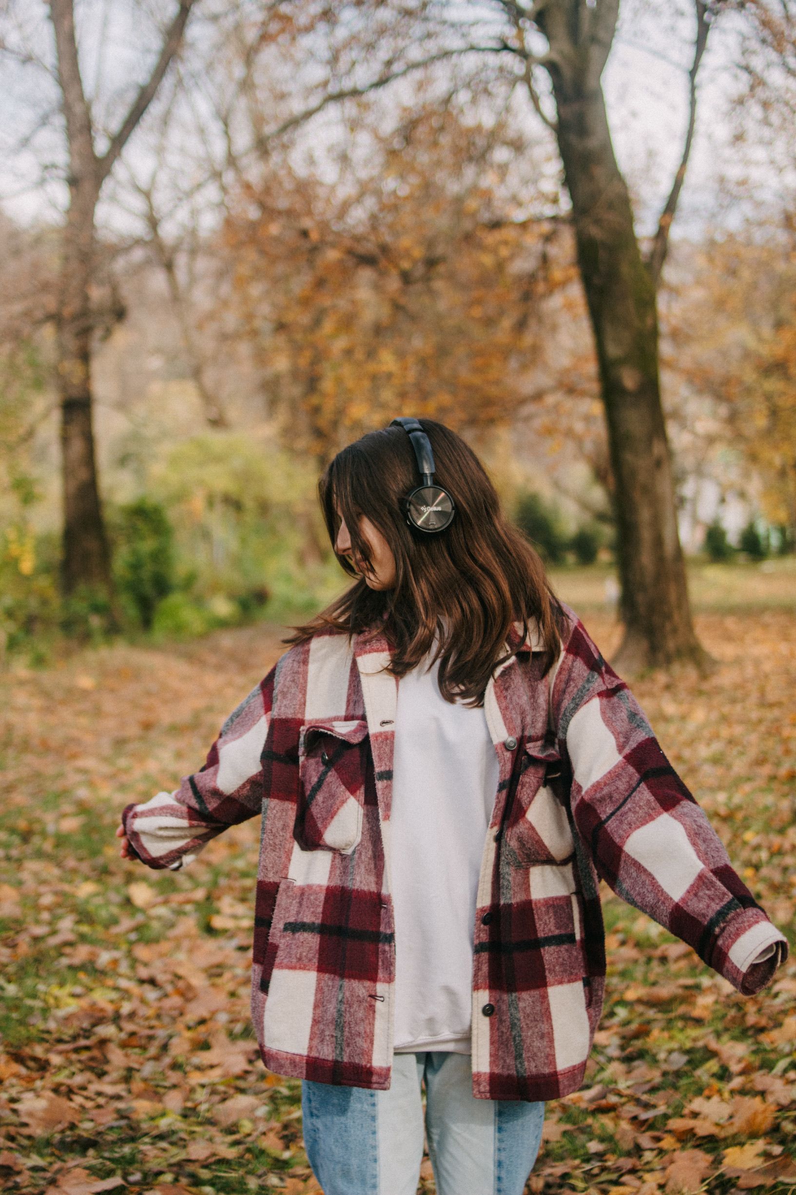 A woman in a checkered jacket and headphones enjoying music while standing in a leafy autumn forest.