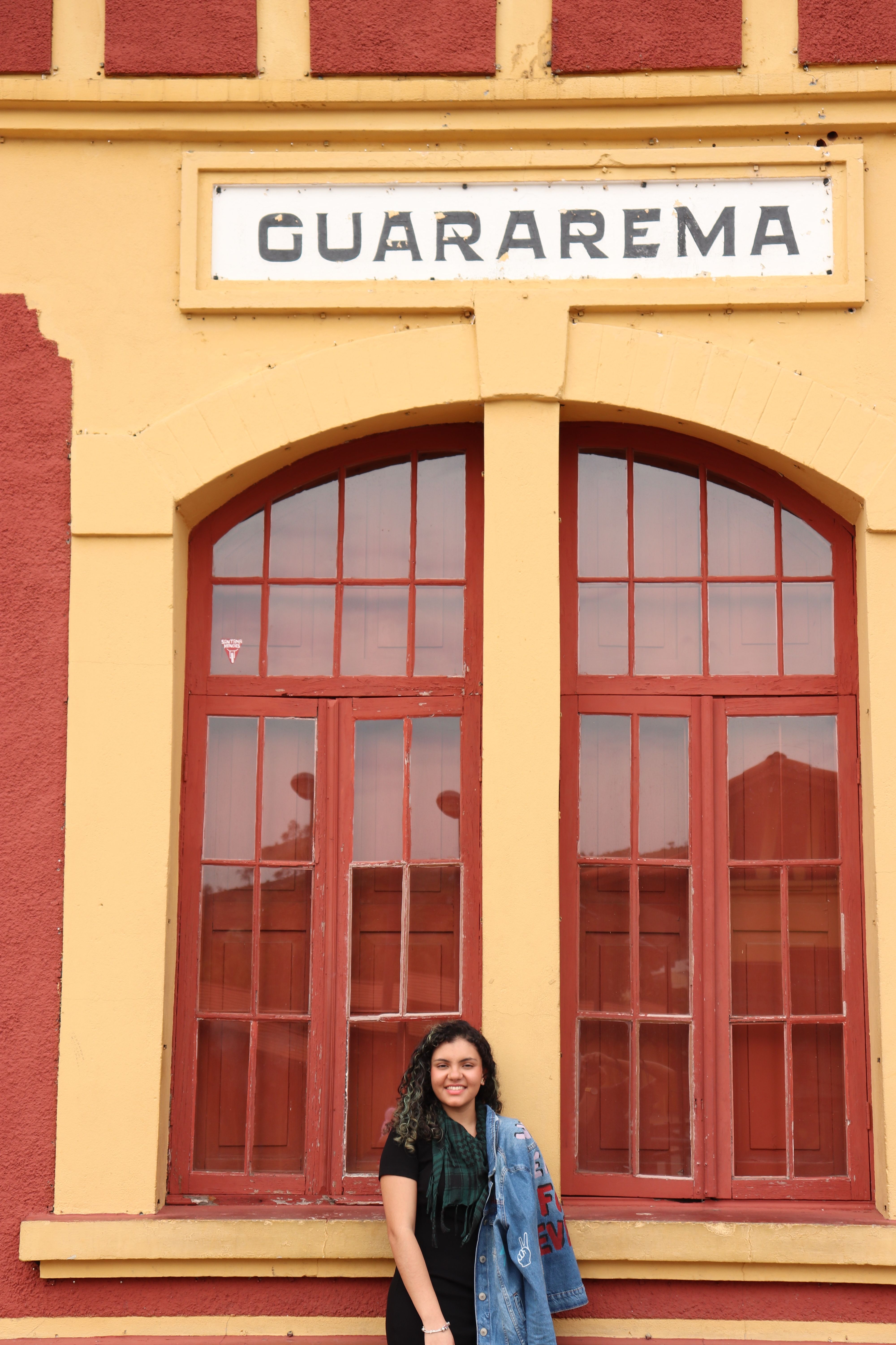 A smiling woman stands outside Guararema railway station in Brazil, framed by red windows.
