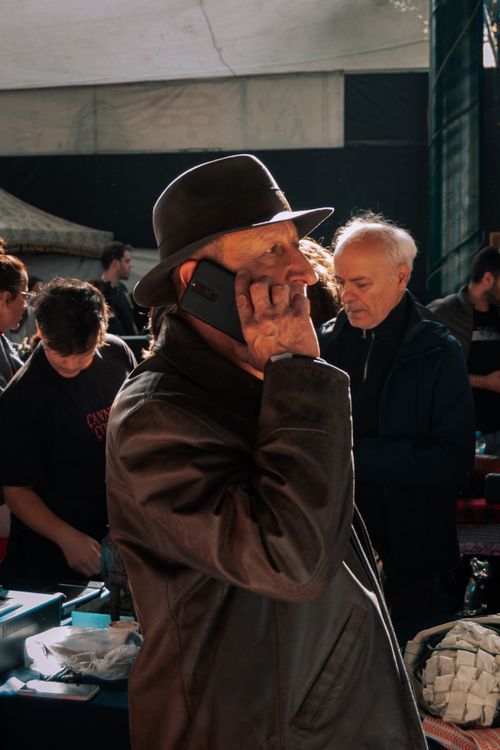 Free Elderly man in a hat talking on smartphone indoors at a marketplace, highlighted by indoor lighting. Stock Photo