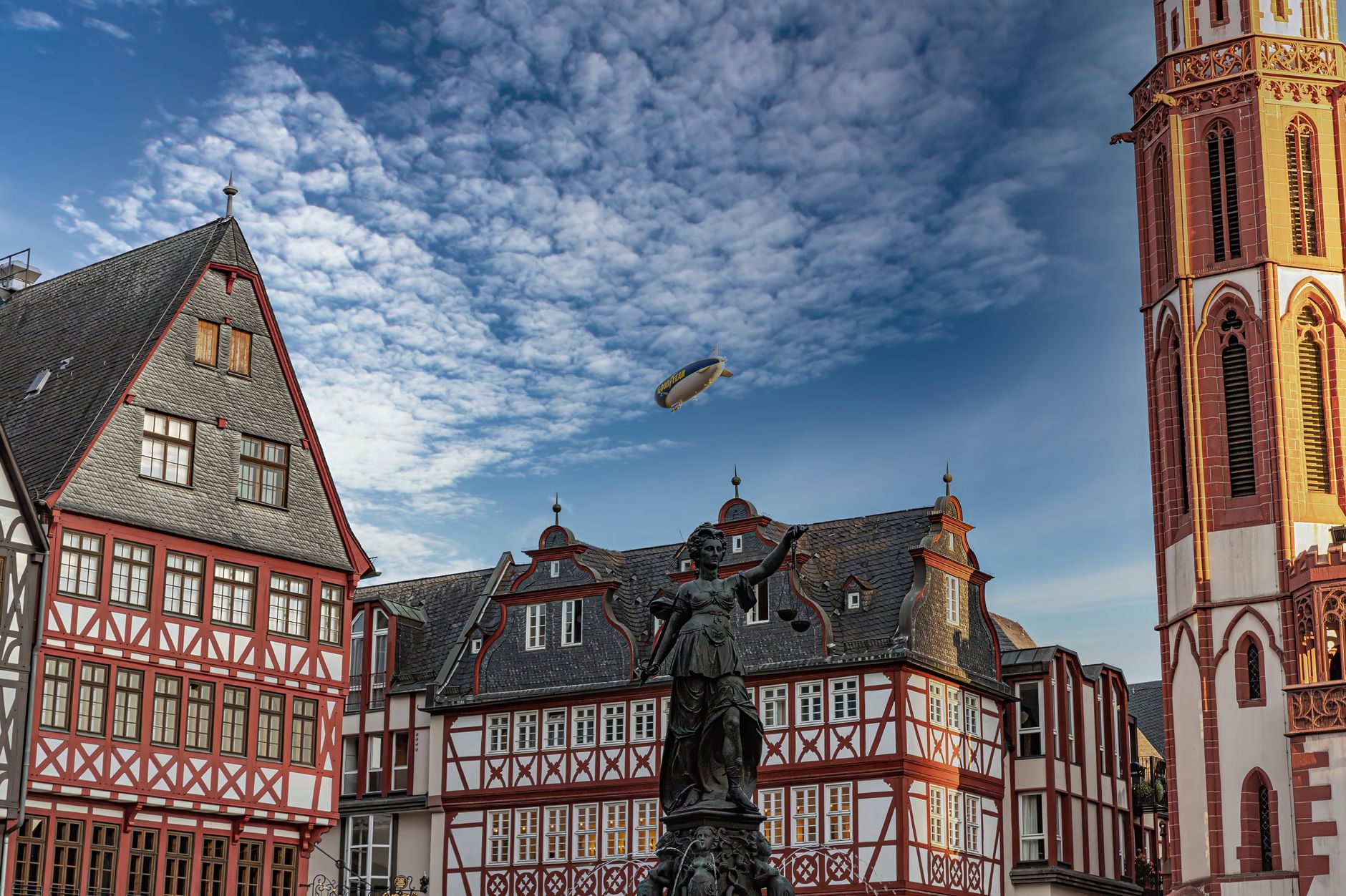 Traditional European architecture with an airship in the sky above a town square.