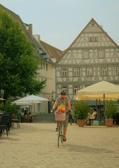 Free A woman rides her bicycle through a charming European town square, enjoying a sunny day. Stock Photo