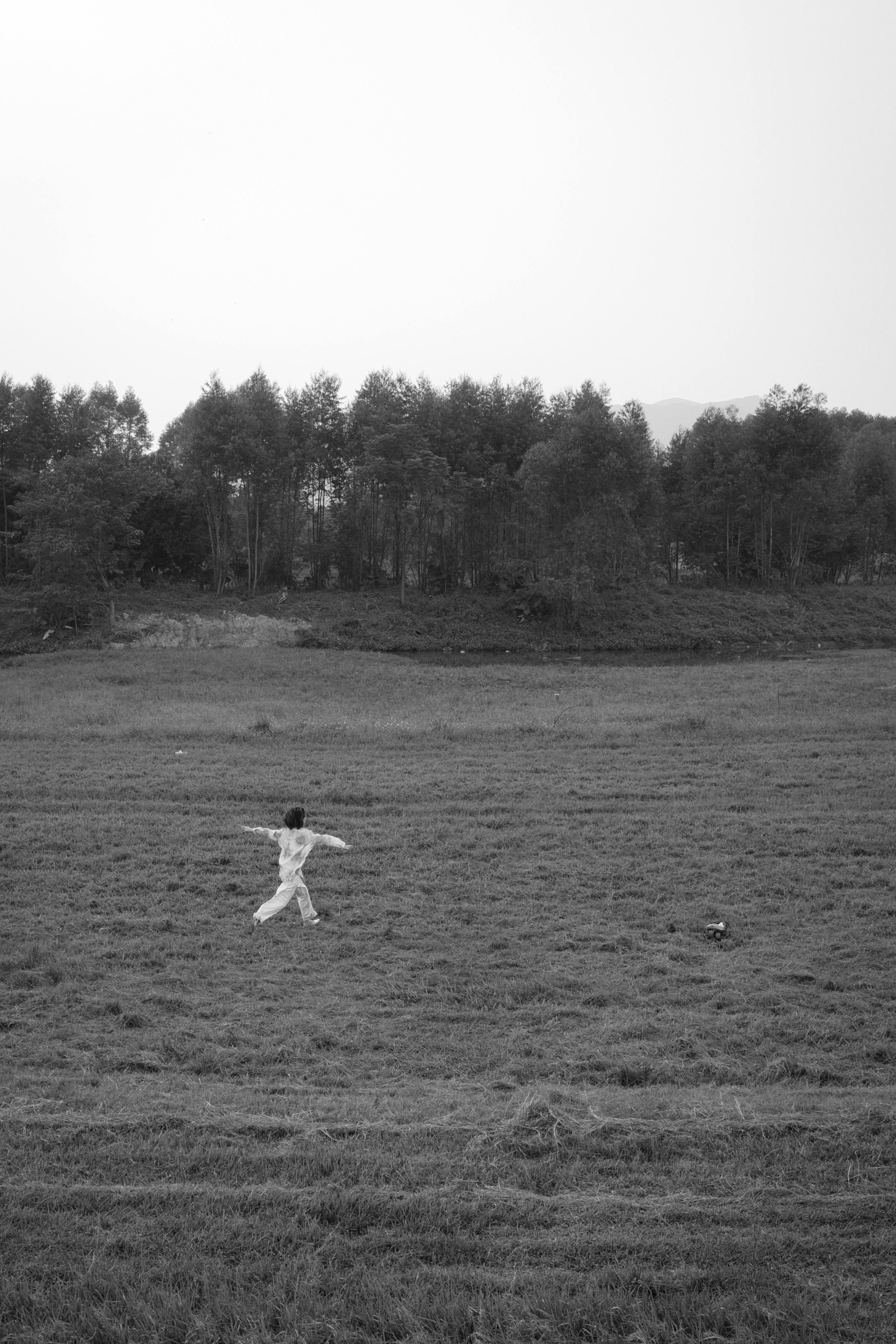 A child joyfully running across a vast field surrounded by trees in black and white.