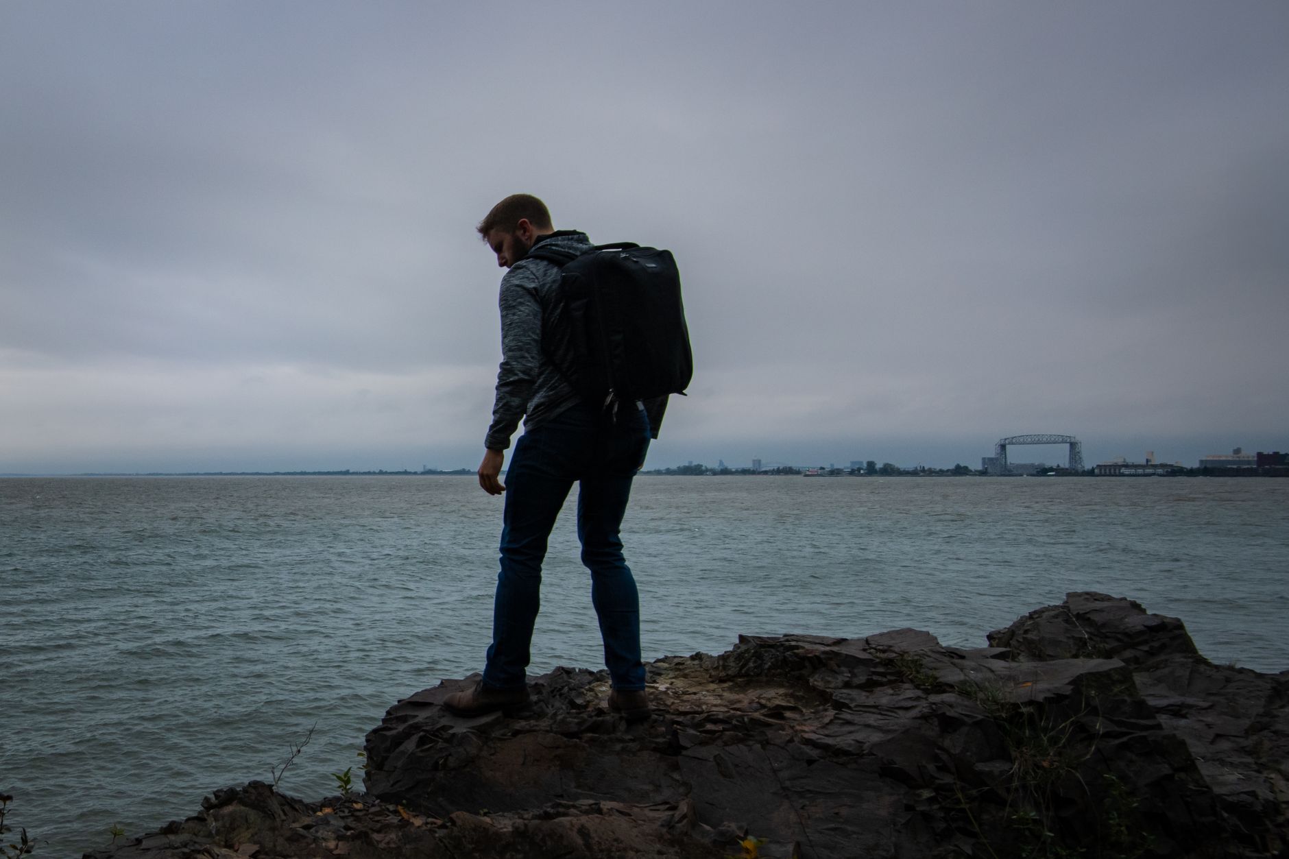 A man with a backpack stands on rocky shore, gazing over Duluth's waterfront.