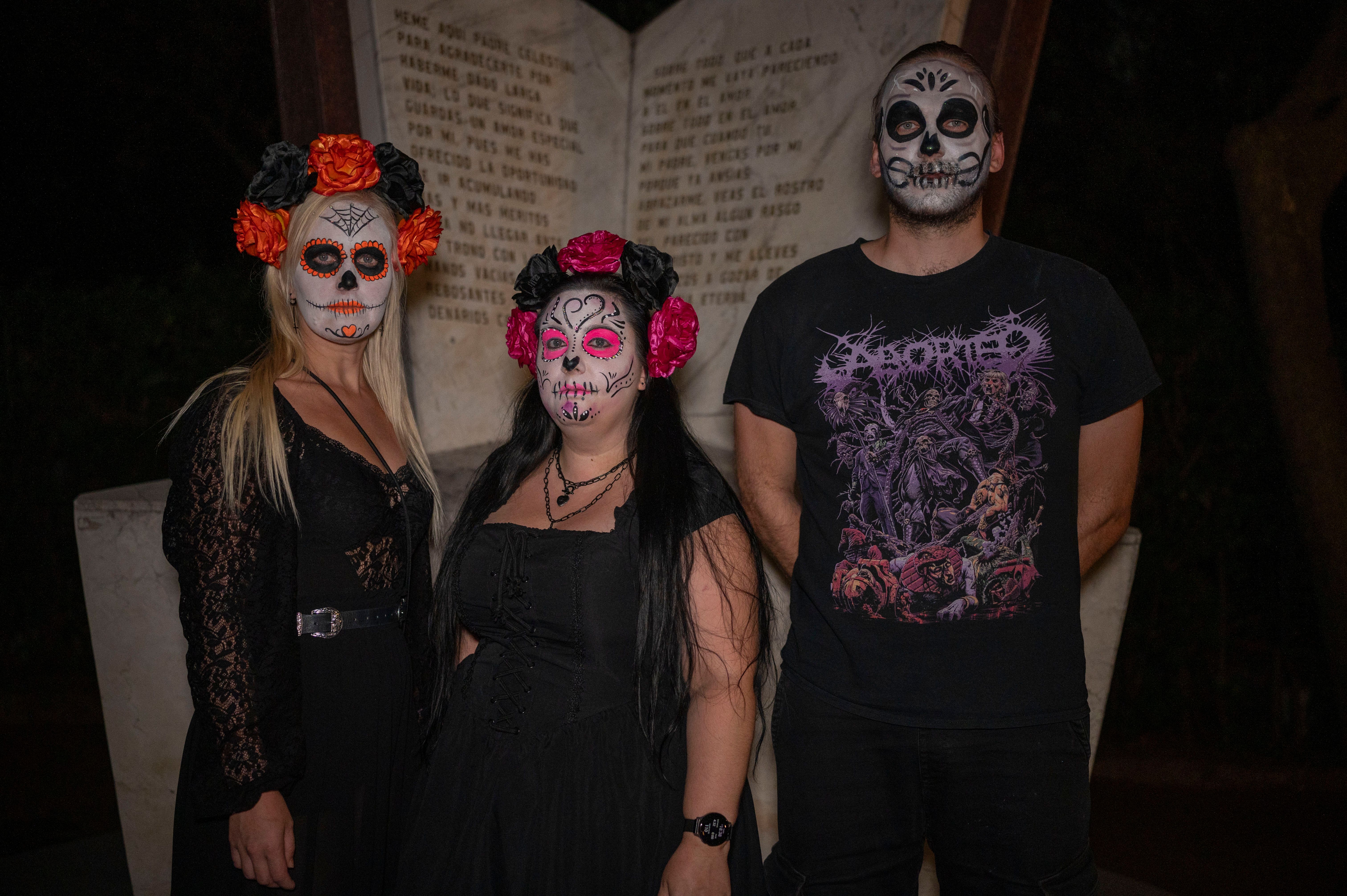 Three people with Day of the Dead makeup pose in front of a monument at night.