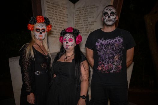 Three people with Day of the Dead makeup pose in front of a monument at night.