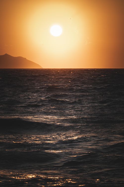 Free Captivating sunset over the ocean with a silhouetted island, waves, and a warm glow. Stock Photo