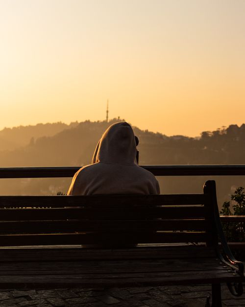 Free A solitary figure in a hoodie sits on a bench, overlooking a serene urban sunset. Stock Photo