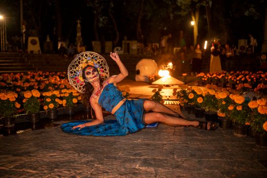 Woman dressed as La Catrina celebrates Day of the Dead with flowers in a candlelit cemetery.