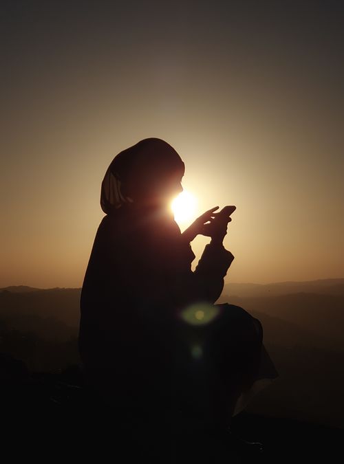 Free Silhouette of a woman in hijab using a smartphone against a striking sunset backdrop. Stock Photo