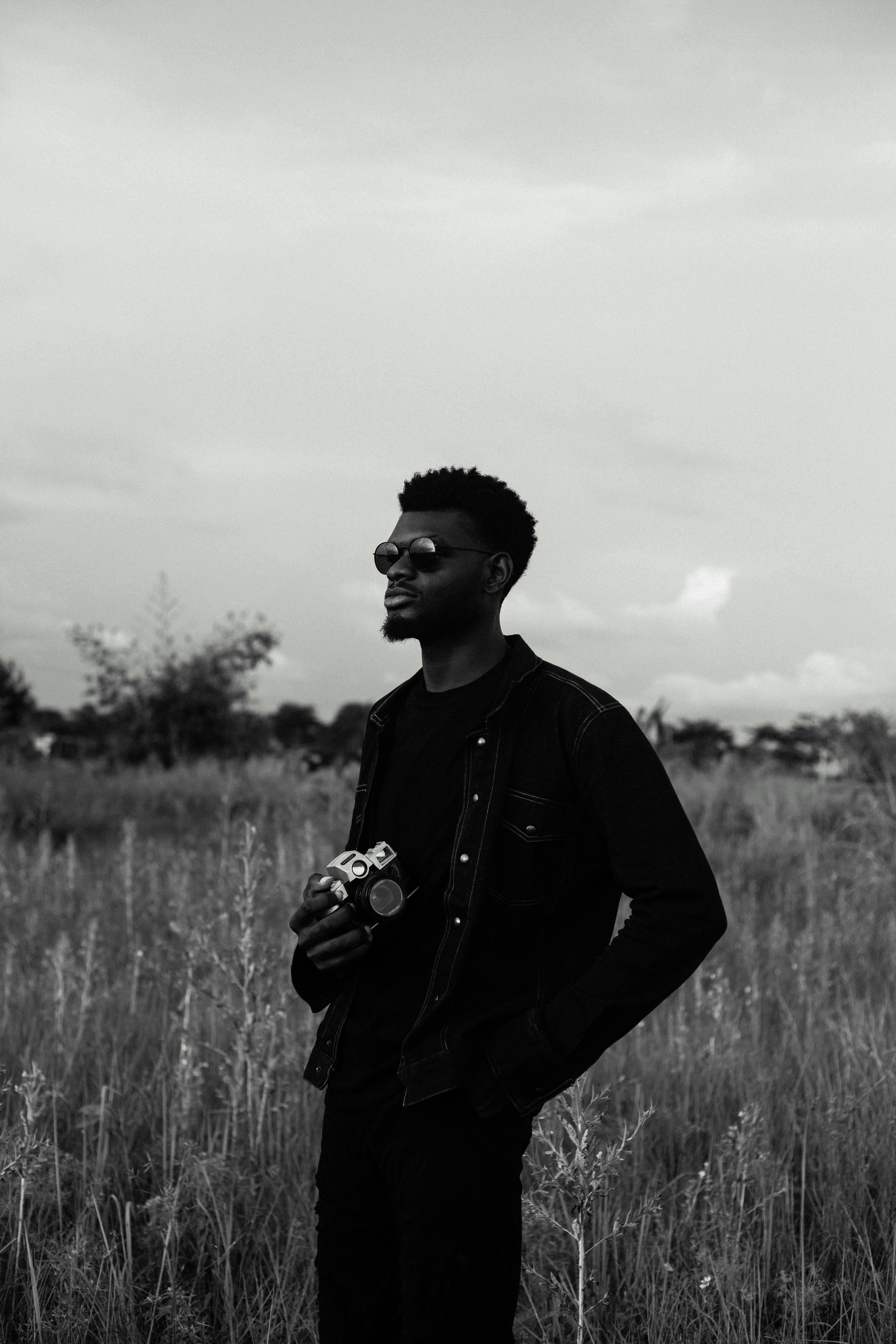 Monochrome portrait of a stylish man with sunglasses holding a camera in a rural field.