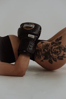 Close-up of a woman's torso with boxing gloves and a detailed floral tattoo on faded background.