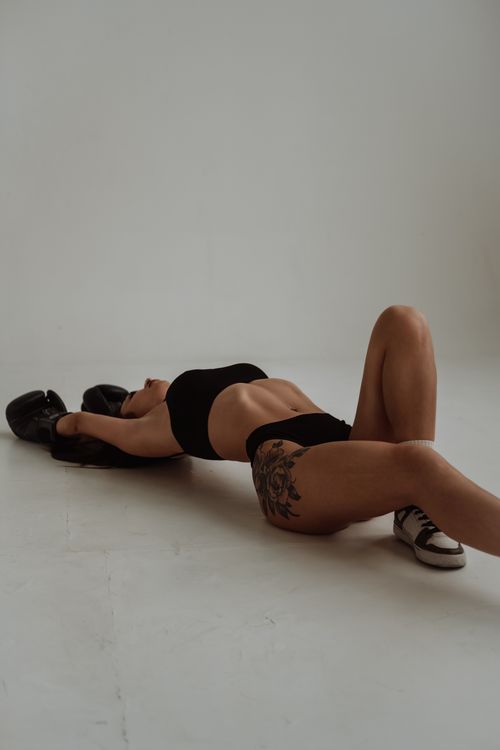 Free Athletic woman lying on studio floor in black bikini with boxing gloves and tattoo. Stock Photo