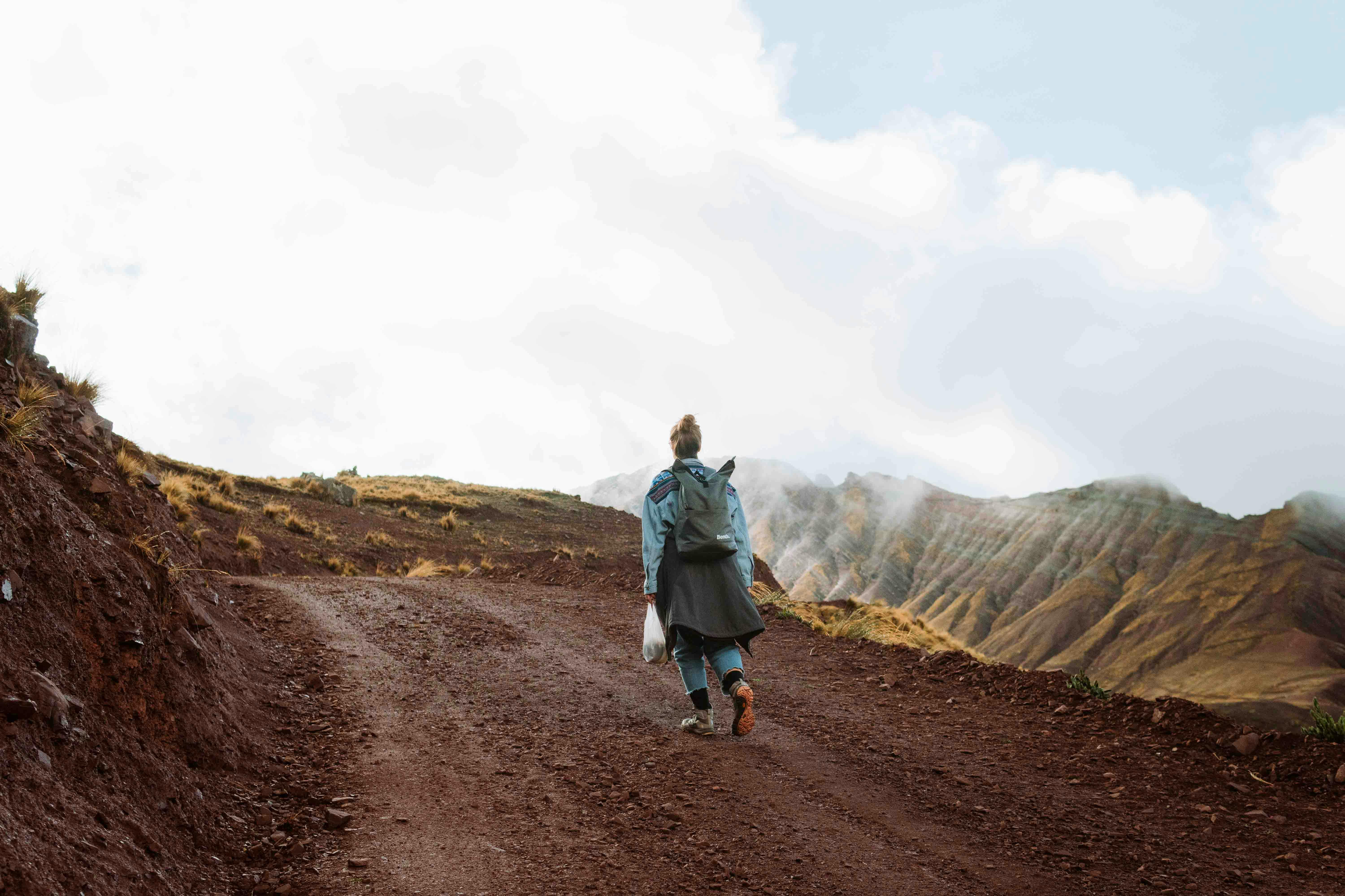 A woman with backpack hiking on a dirt path in a rugged mountain landscape, under a cloudy sky.