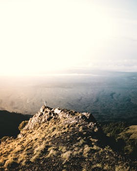 Solitary hiker stands on a rugged mountain peak at sunrise, overlooking a vast landscape.