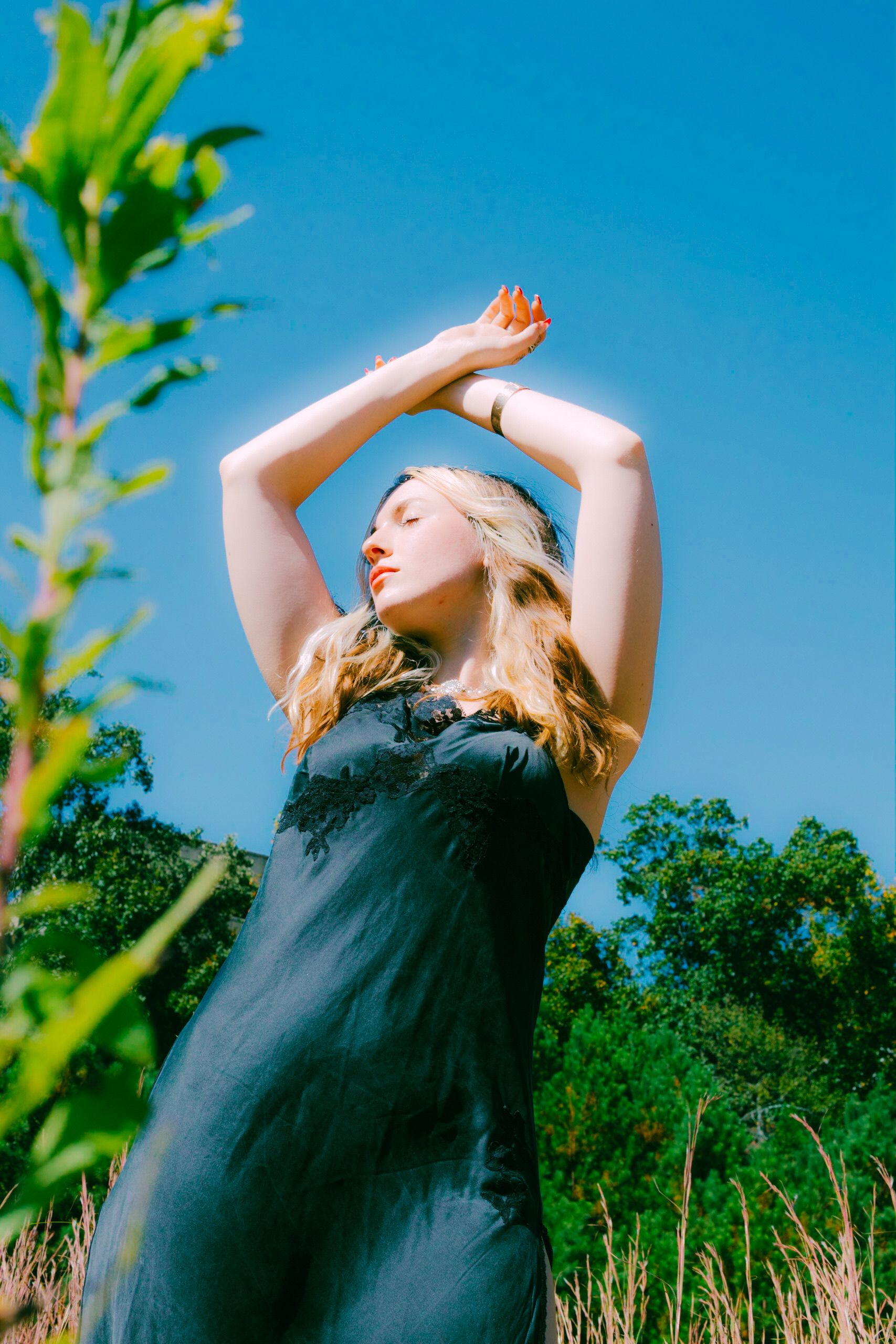 Woman in black dress posing with arms raised against clear blue sky outdoors.