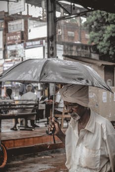 Elderly man with turban holds an umbrella, walking through a bustling city street in the rain.