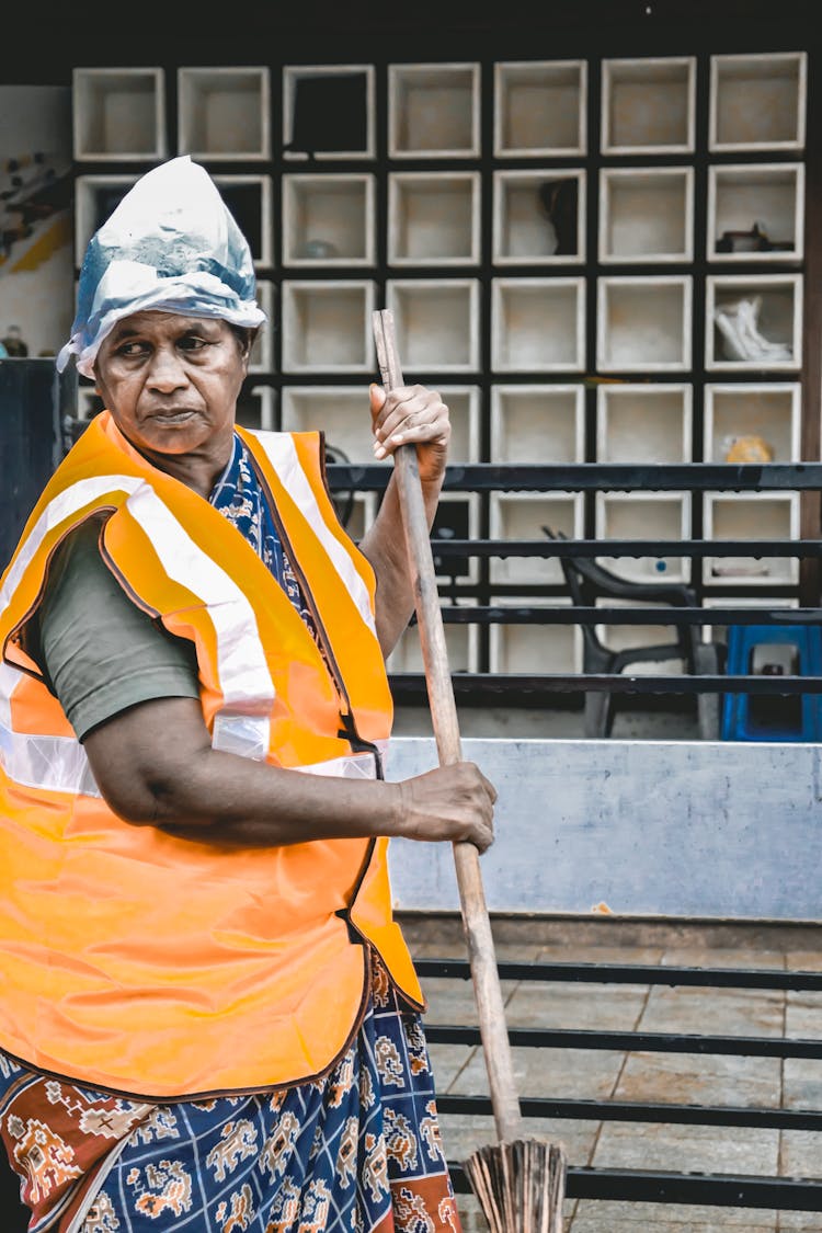 Elderly Woman In A Reflective Vest Sweeping The Street 