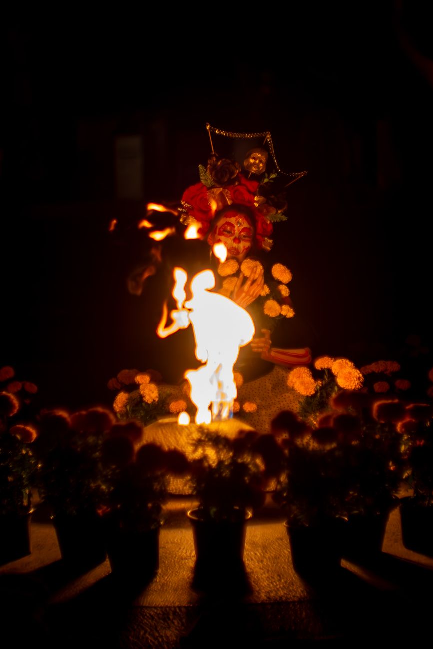 A woman with traditional Día de los Muertos makeup celebrates at night with candles and decorations.
